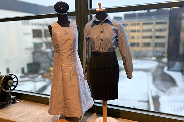 Two mannequins display vintage clothing by a large window. The left mannequin wears a light-colored sleeveless dress with embroidery, while the right one sports a blue collared shirt and dark skirt. Snowy urban landscape visible outside.
