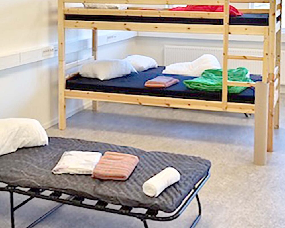 En sovsal med en våningssäng i trä i bakgrunden och en tältsäng med mörk madrass i förgrunden. Kuddar och vikta lakan placeras på varje säng. Soligt utomhusljus syns genom fönstren.