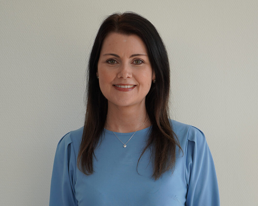 Kvinna med långt mörkt hår klädd i en ljusblå blus, står mot en ljus bakgrund. Hon ler vänligt och bär ett subtilt smycke runt halsen. Hennes uttryck är professionellt och vänligt.