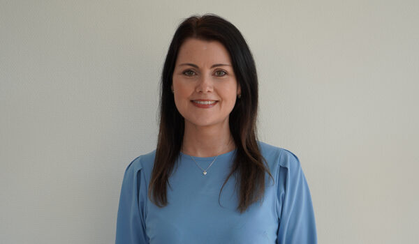 Kvinna med långt mörkt hår klädd i en ljusblå blus, står mot en ljus bakgrund. Hon ler vänligt och bär ett subtilt smycke runt halsen. Hennes uttryck är professionellt och vänligt.