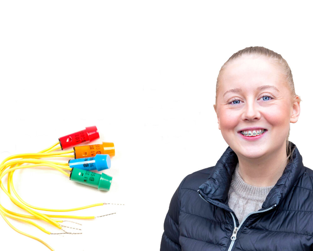 En kvinna med tandställning ler mot kameran, klädd i en svart jacka. Till vänster finns flerfärgade elektronikkablar med blå, röda, gröna och gula kontakter. Vit bakgrund med fokus på teknik och personligt uttryck.