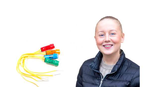 En kvinna med tandställning ler mot kameran, klädd i en svart jacka. Till vänster finns flerfärgade elektronikkablar med blå, röda, gröna och gula kontakter. Vit bakgrund med fokus på teknik och personligt uttryck.