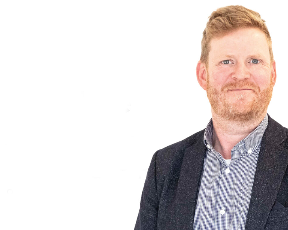 En man med kort hår och skägg bär en mörk kavaj och en rutig skjorta. Han står mot en vit bakgrund och ler lätt. Fotografiet verkar vara taget för professionella ändamål, exempelvis ett nyhetsbrev.