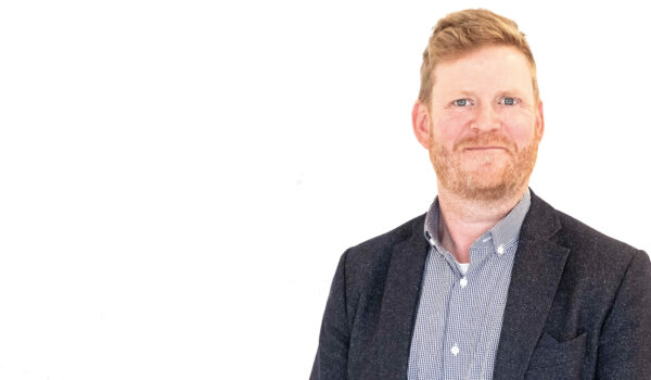 En man med kort hår och skägg bär en mörk kavaj och en rutig skjorta. Han står mot en vit bakgrund och ler lätt. Fotografiet verkar vara taget för professionella ändamål, exempelvis ett nyhetsbrev.