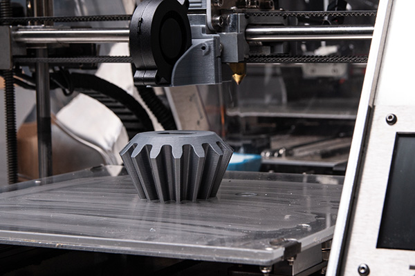 A 3D printer fabricates a gray, gear-shaped object on its build platform. The printer's nozzle, partially visible, deposits material layer by layer. The surrounding machinery features metal and plastic components, indicating an industrial setting.