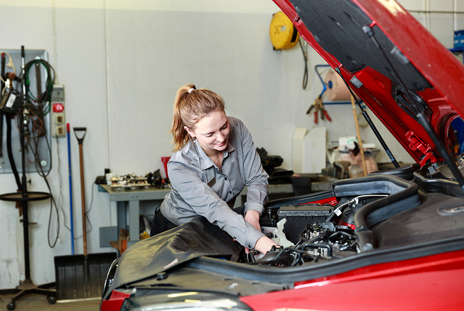 Mechanic working under the hood of a red car in a workshop, wearing a gray shirt and bending over to adjust engine components. Workshop background includes tools, organized equipment, and shelves with automotive parts.