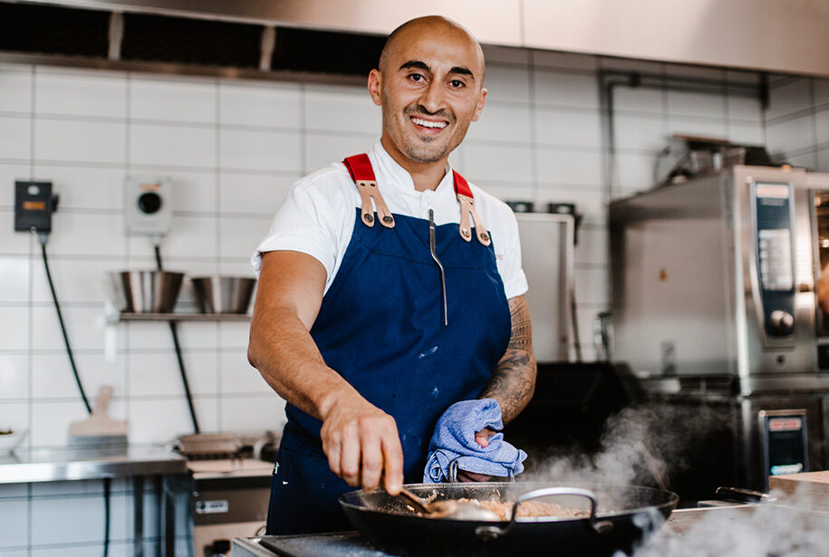 Chef wearing a blue apron smiles while cooking in a professional kitchen. He stirs a steaming pan with a spoon, holding a blue cloth. The kitchen has white tiled walls and stainless steel appliances in the background.