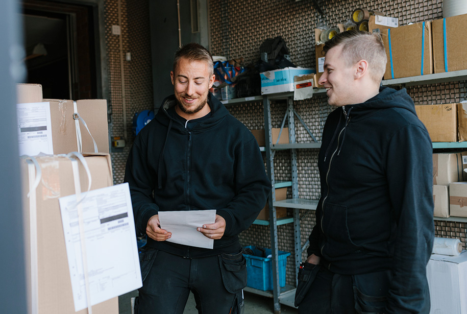 Two people in black hoodies stand in a storage room, smiling at each other. One holds a document, possibly discussing package details. Cardboard boxes with labels are stacked nearby, and shelves filled with various items line the wall.