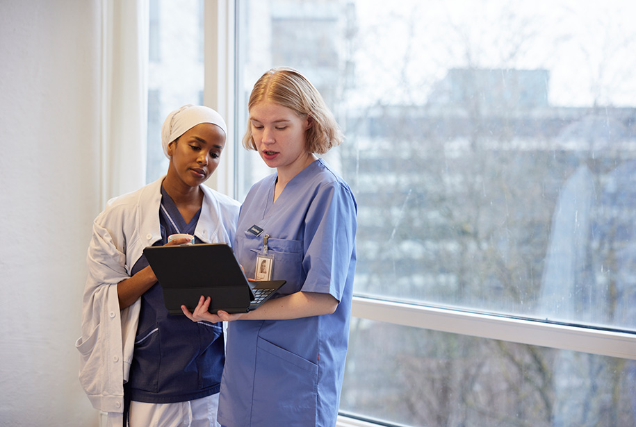 Two medical professionals in scrubs and lab coats discuss patient information while looking at a laptop. They stand near a large window with a blurred view of the outside, indicating a hospital setting.